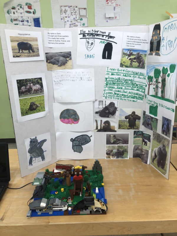 A LEGO Robotics Expo Display. A trifold contains text describing and images showing hippopotamuses and gorillas. In front of the trifold there is a Jungle made with LEGO, with a monkey hippopotamus built with LEGO.