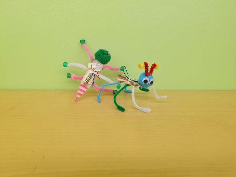 Two Jitterbugs on a table, made of popsicle sticks, pompoms, googly eyes, and pipecleaners. They have small motors connected via wires to coin cell batteries.