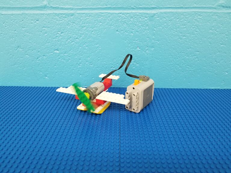 A lego WeDo Plane on a blue background. The green plane propeller is blurred in motion.